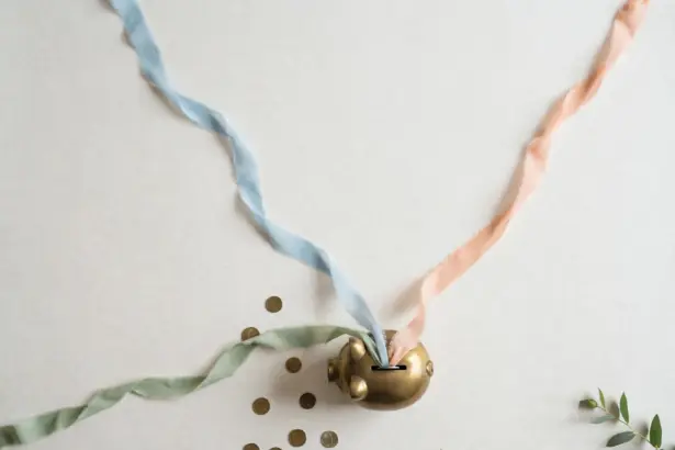 Flat-lay: three thin pastel ribbons in blue, peach and sage streaming across an off-white background and converging into a small round brass piggy bank, with scattered coins and a green leaf at the edge