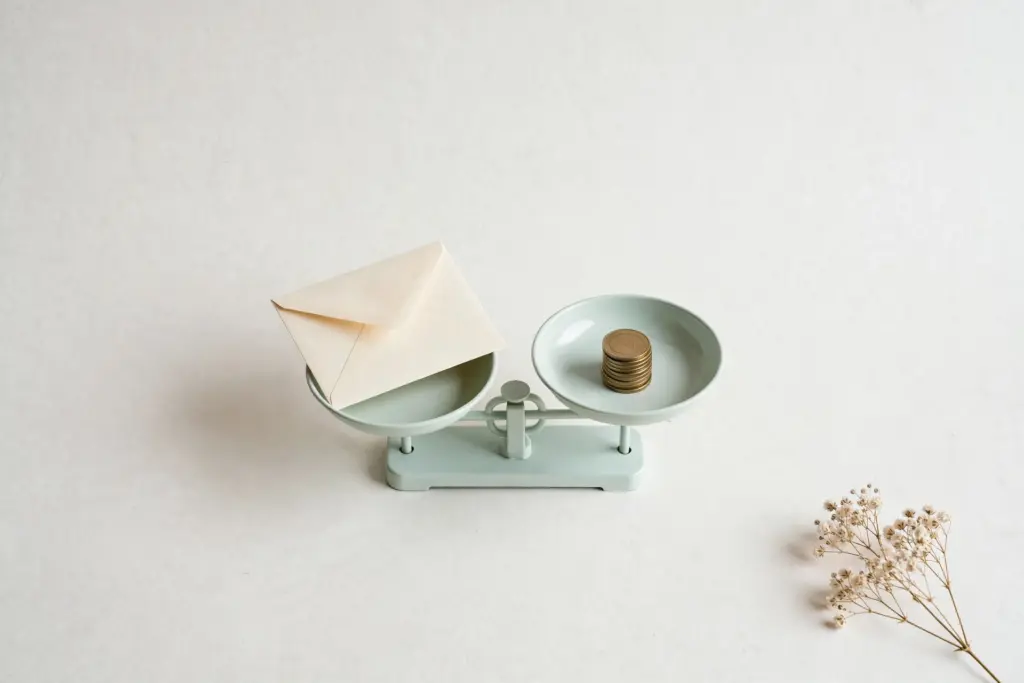 Flat-lay: a pale mint kitchen scale weighing a cream paper envelope on one pan against a short stack of coins on the other, with a dried gypsophila sprig in the corner