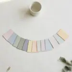 Flat-lay: a single white ceramic cup at the top centre with twelve small pastel paper cards fanned out in an arc below it on an off-white background, a sage ribbon and brass paperclip at the edges