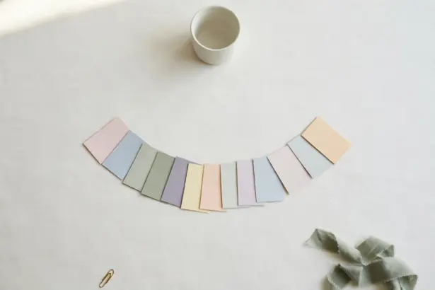 Flat-lay: a single white ceramic cup at the top centre with twelve small pastel paper cards fanned out in an arc below it on an off-white background, a sage ribbon and brass paperclip at the edges