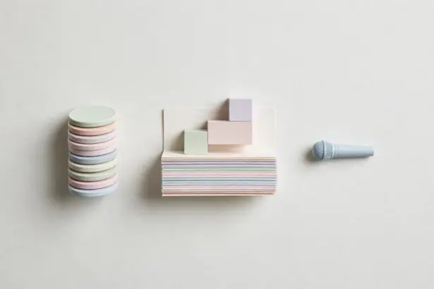 Flat-lay: a tower of stacked pastel coin discs beside layered paper sheets topped with small pastel blocks like a layer cake, and a tiny pale blue microphone to the right