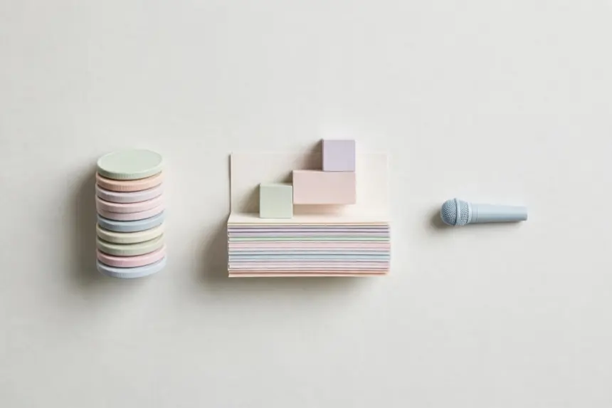 Flat-lay: a tower of stacked pastel coin discs beside layered paper sheets topped with small pastel blocks like a layer cake, and a tiny pale blue microphone to the right