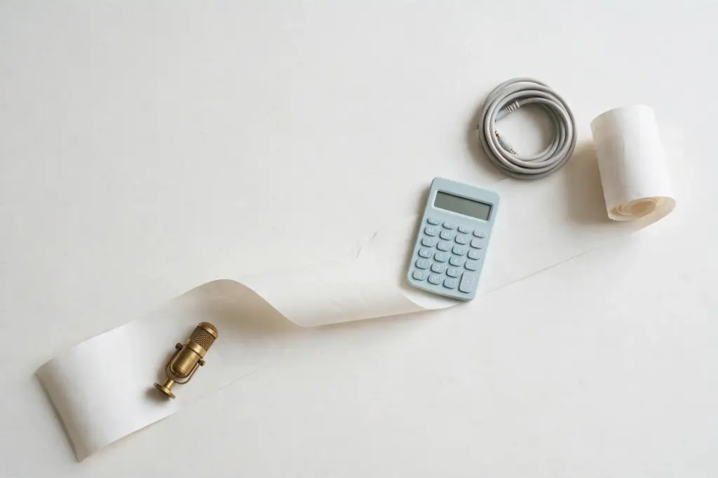 Flat-lay: a long pale paper receipt unrolled and curling across an off-white desk, held down by a small brass microphone, a pastel blue calculator and a coiled grey audio cable