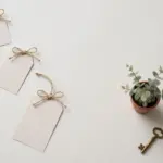 Flat-lay: three blank paper price tags of increasing size tied with twine bows in a diagonal row, beside a small potted eucalyptus plant and a vintage brass key