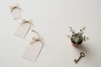 Flat-lay: three blank paper price tags of increasing size tied with twine bows in a diagonal row, beside a small potted eucalyptus plant and a vintage brass key