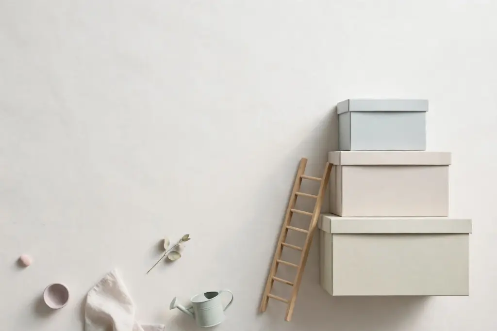 Flat-lay: a small wooden ladder leaning against a stack of three pale storage boxes of increasing height on an off-white background, with a tiny pale watering can at the base
