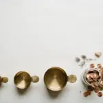 Flat-lay: three brass measuring cups of increasing size lined up in a row beside a small glass jar overflowing with copper coins on an off-white background