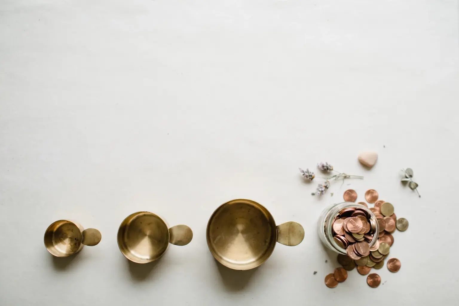 Flat-lay: three brass measuring cups of increasing size lined up in a row beside a small glass jar overflowing with copper coins on an off-white background