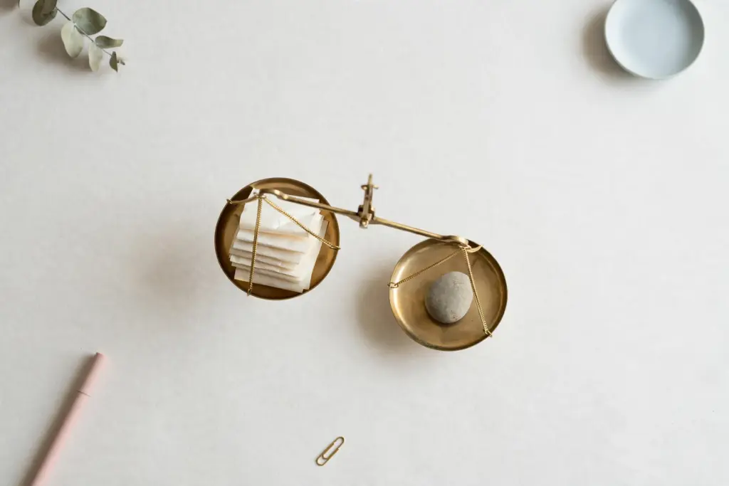 Flat-lay: a small brass balance scale weighing a stack of paper receipts against a single smooth stone, the stone side sitting lower, on a pale off-white desk