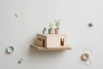 Flat-lay: a small wooden house model on a single flat base holding three tiny potted plants of different heights on its flat roof, on a pale off-white background