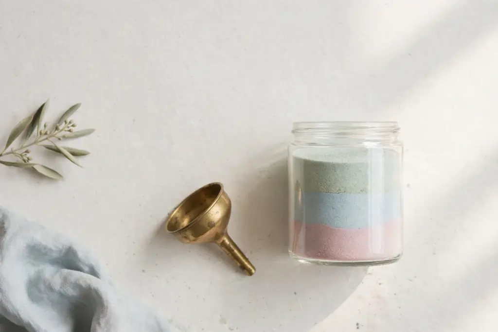 Flat-lay: a clear glass jar filled with three bands of layered pastel sand in pink, blue and sage beside a small brass funnel on an off-white desk with a soft blue cloth