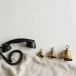 Flat-lay: a black vintage rotary telephone handset with a coiled cord resting on a linen cloth beside three brass bells of increasing size