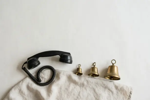 Flat-lay: a black vintage rotary telephone handset with a coiled cord resting on a linen cloth beside three brass bells of increasing size