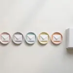 Flat-lay: a neat row of six small white envelopes each resting on its own pastel saucer, with a larger stack of white envelopes waiting beside them