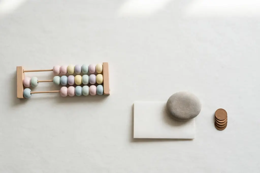 Flat-lay: a wooden abacus with pastel beads pushed mostly to one side, next to a blank paper sheet held down by a smooth stone paperweight and a small stack of coins