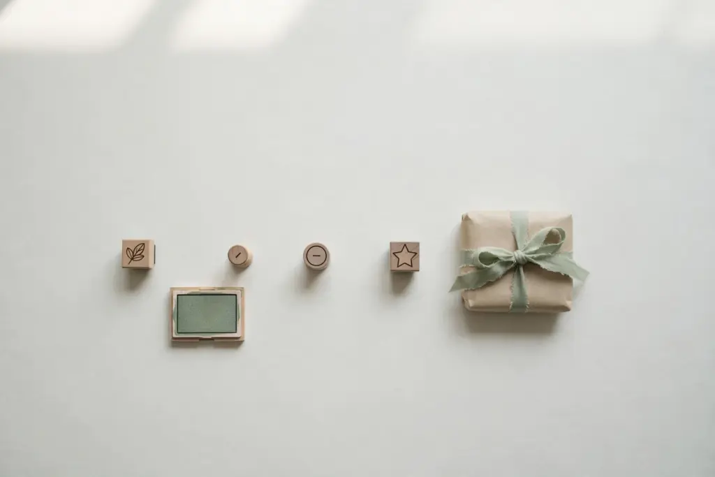 Flat-lay: a finished paper parcel tied with sage ribbon at the end of a short row of small wooden stamps and a green ink pad on a pale off-white desk
