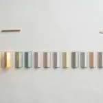Flat-lay: fifteen small paper doors in a row on a pale off-white background, the nearest ones open with warm light spilling out and the far ones still closed