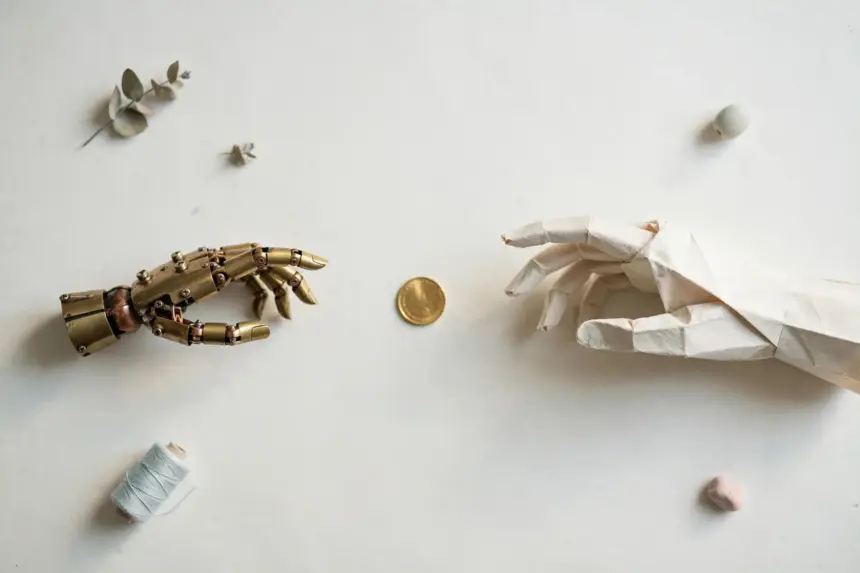 Flat-lay: a small brass robot hand and a paper hand reaching toward the same gold coin from opposite sides on a pale off-white background