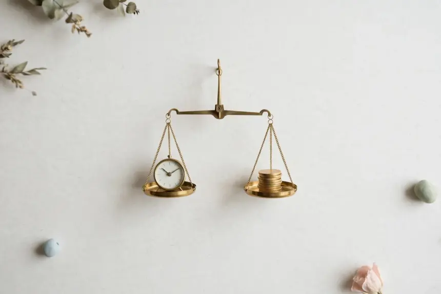 Flat-lay: a brass balance scale on a pale off-white background with a small clock on one pan and a neat stack of gold coins on the other, perfectly level