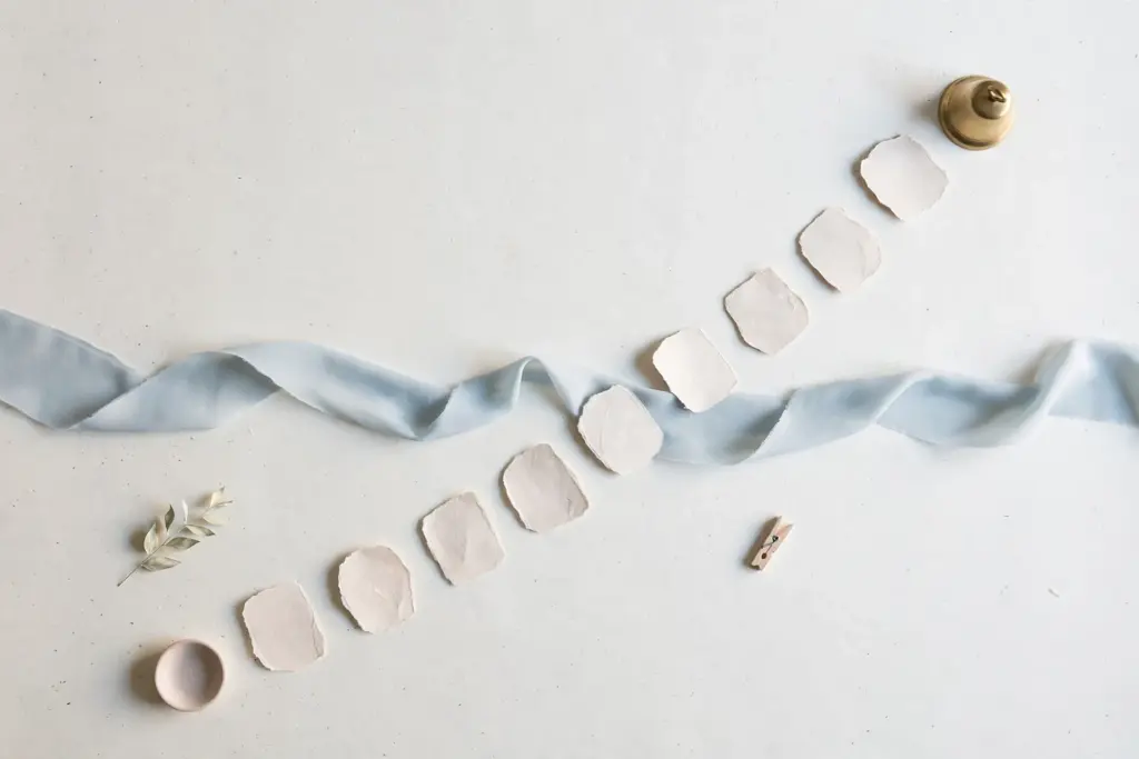 Flat-lay: nine small paper stepping stones crossing a pale stream of blue ribbon on an off-white desk toward a small brass bell