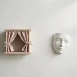 Flat-lay: a small wooden puppet stage with a tiny parted curtain on a pale off-white background, a plain white ceramic mask resting beside it
