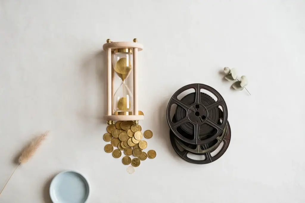 Flat-lay: an hourglass standing next to a neat stack of vintage film reels on a pale off-white desk, with a small pile of gold coins beside them