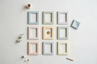 Flat-lay: a neat three-by-three grid of small pastel picture frames on a pale off-white background, the center frame holding a single gold coin instead of a picture