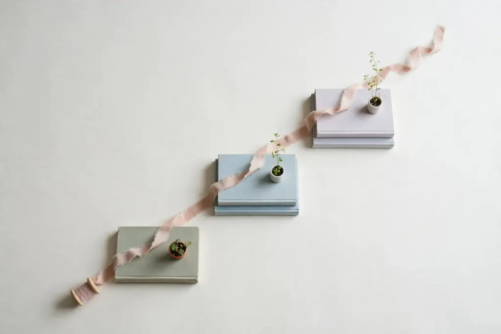 Flat-lay: a small staircase of stacked books on a pale off-white desk with a tiny potted plant growing taller on each step and a spool of ribbon unspooling up the steps