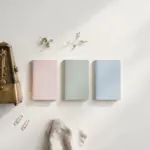 Flat-lay: a vintage brass metronome beside a neat row of small pastel notebooks on a pale off-white background