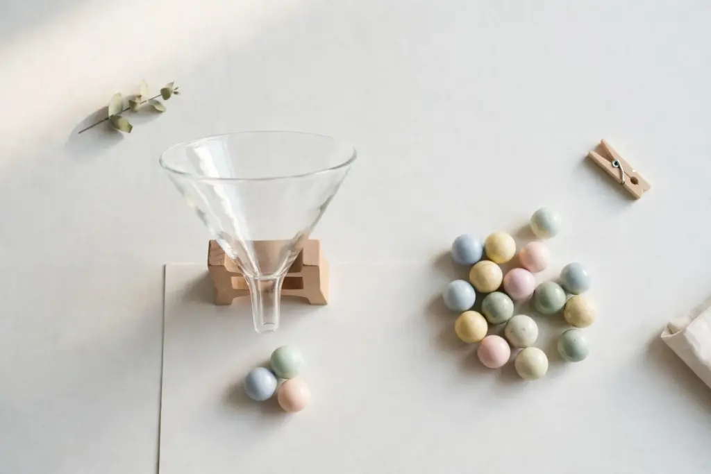 Flat-lay: a clear glass funnel resting on a wooden stand on a pale off-white desk, three smooth pastel marbles passing through it and a larger scattered pile waiting beside it