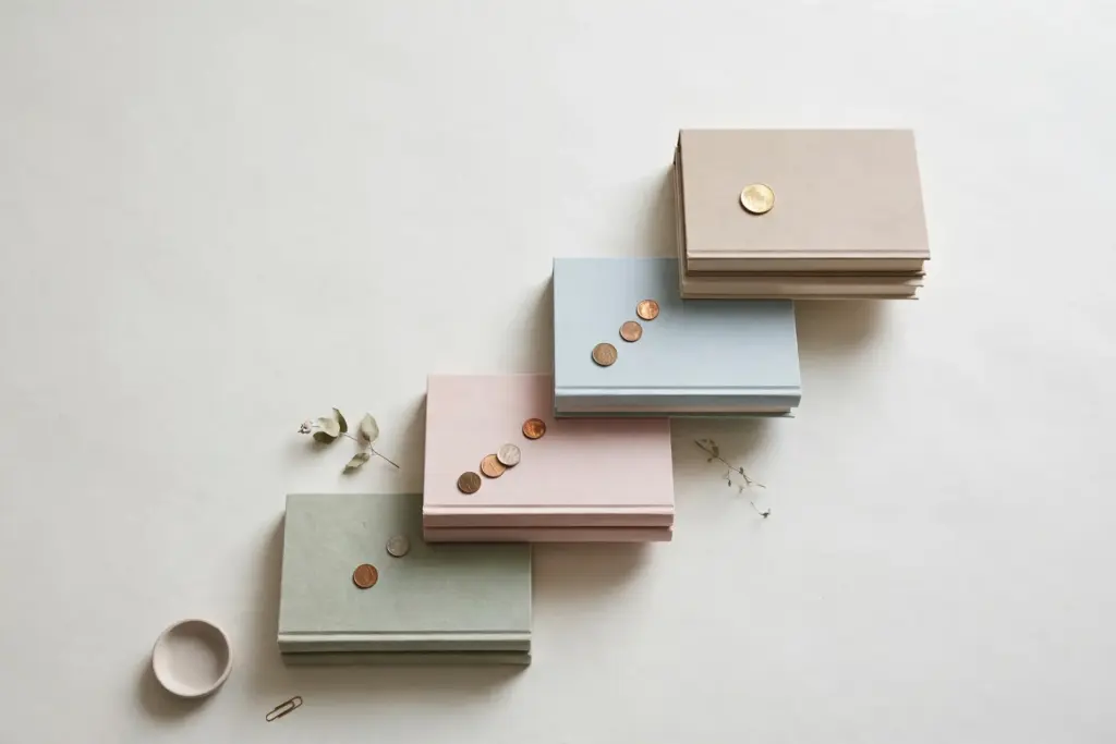 Flat-lay: a gently rising staircase made of stacked hardcover books on a pale off-white desk, a single gold coin resting on the top step and a trail of smaller coins on the steps below
