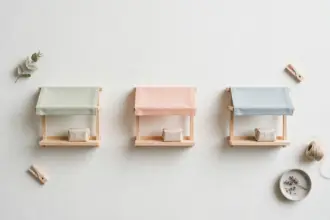 Flat-lay: three miniature market stalls in pastel canvas and wood standing in a row on a pale off-white background, each with one small blank paper parcel on its counter