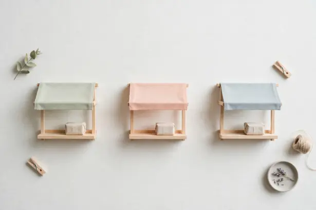 Flat-lay: three miniature market stalls in pastel canvas and wood standing in a row on a pale off-white background, each with one small blank paper parcel on its counter