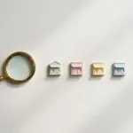 Flat-lay: a small brass magnifying glass resting beside a neat row of tiny pastel shopfront models on a pale off-white background