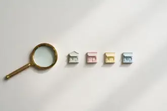 Flat-lay: a small brass magnifying glass resting beside a neat row of tiny pastel shopfront models on a pale off-white background