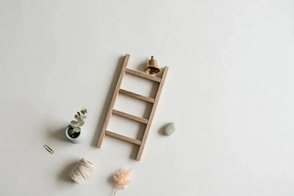 Flat-lay: a wooden ladder with four wide rungs leaning on a pale off-white desk, a small brass bell resting on its top rung
