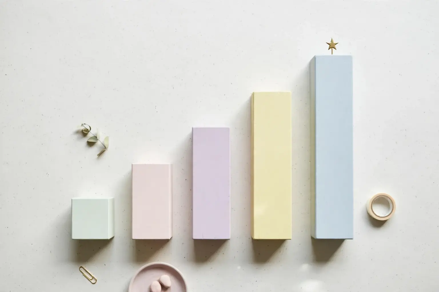 Flat-lay: a row of small ascending bar-chart blocks in pastel wood on a pale off-white background, the tallest block topped with a tiny brass star