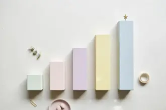 Flat-lay: a row of small ascending bar-chart blocks in pastel wood on a pale off-white background, the tallest block topped with a tiny brass star