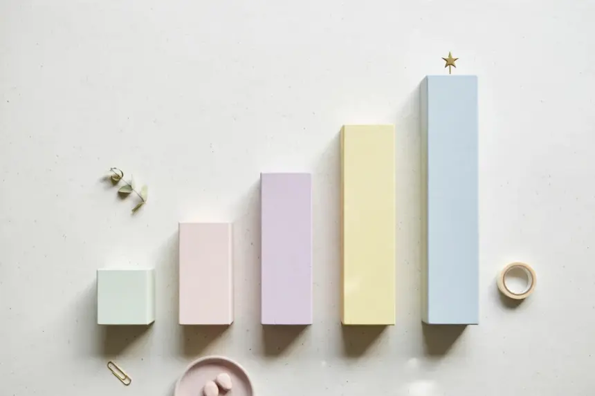 Flat-lay: a row of small ascending bar-chart blocks in pastel wood on a pale off-white background, the tallest block topped with a tiny brass star