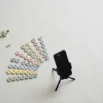 Flat-lay: a smartphone on a small tripod surrounded by a fan of twenty tiny pastel paper play-buttons on a pale off-white background