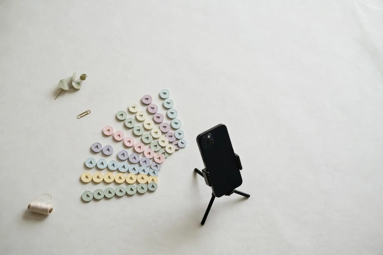 Flat-lay: a smartphone on a small tripod surrounded by a fan of twenty tiny pastel paper play-buttons on a pale off-white background