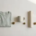 Flat-lay: three blank products in a row on a pale off-white background — a folded plain tee, a plain ceramic mug and a rolled paper poster — each with a small brass price-weight beside it