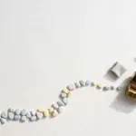 Flat-lay: a winding path of small pastel stepping stones, the first ones small and close together, leading across a pale off-white desk toward a tiny brass treasure chest left ajar