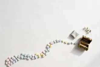 Flat-lay: a winding path of small pastel stepping stones, the first ones small and close together, leading across a pale off-white desk toward a tiny brass treasure chest left ajar