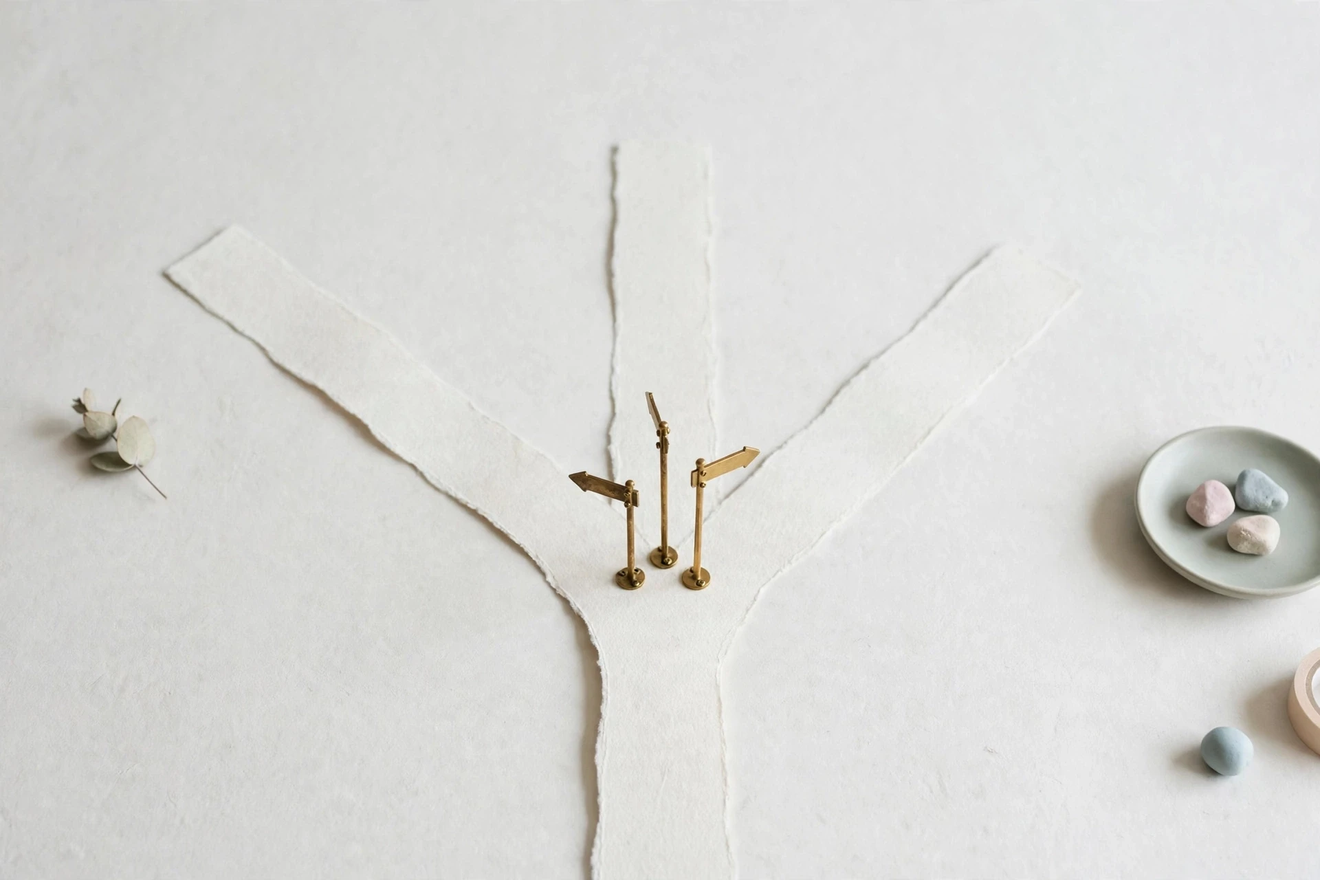 Flat-lay: three small brass signposts of different heights standing at a fork on a pale paper road, each pointing a different direction, on an off-white background