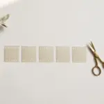 Flat-lay: one long ribbon of pale film strip cut into several neat short segments laid in a row, with small brass scissors resting beside them on a pale off-white background