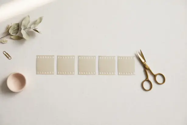 Flat-lay: one long ribbon of pale film strip cut into several neat short segments laid in a row, with small brass scissors resting beside them on a pale off-white background