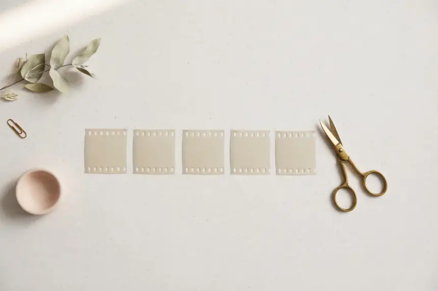 Flat-lay: one long ribbon of pale film strip cut into several neat short segments laid in a row, with small brass scissors resting beside them on a pale off-white background