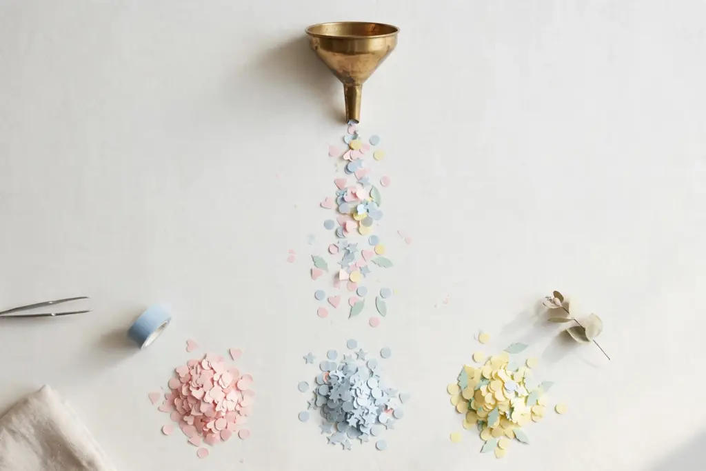 Flat-lay: a small brass funnel pouring a stream of tiny pastel paper shapes into several neat piles sorted by color below it, on a pale off-white desk