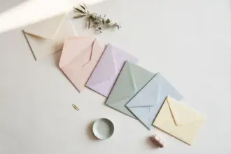 Flat-lay: a single cream envelope at the start of a growing trail of pastel envelopes fanning outward across a pale off-white desk